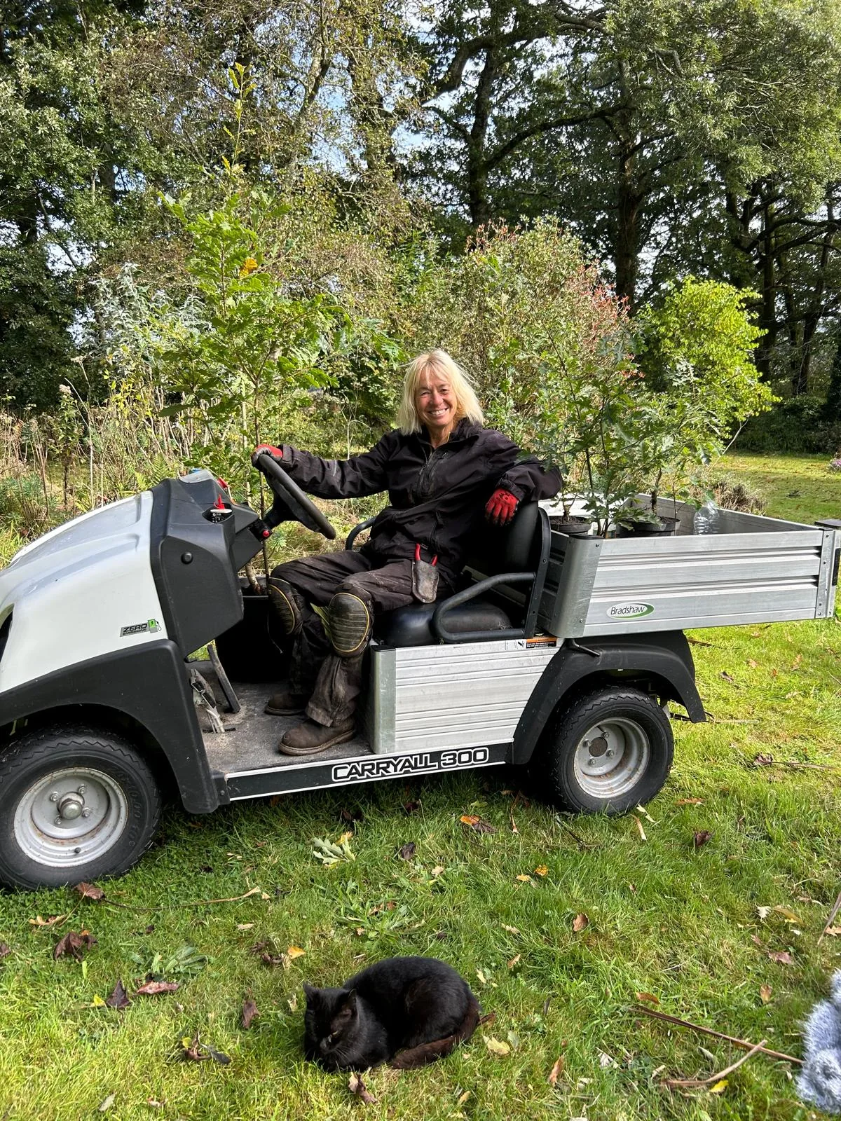 The Curious Garden designer smiling from a Carryall work buggy with a cat on the grass