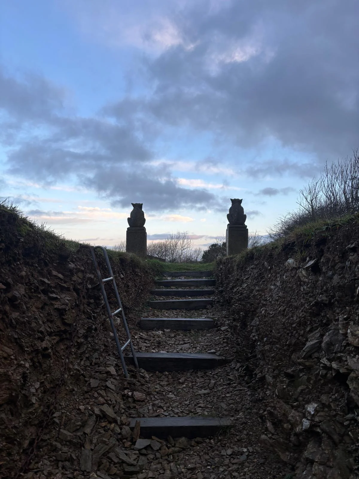 A moody garden pathway rising towards two owl statues beneath a dramatic sky